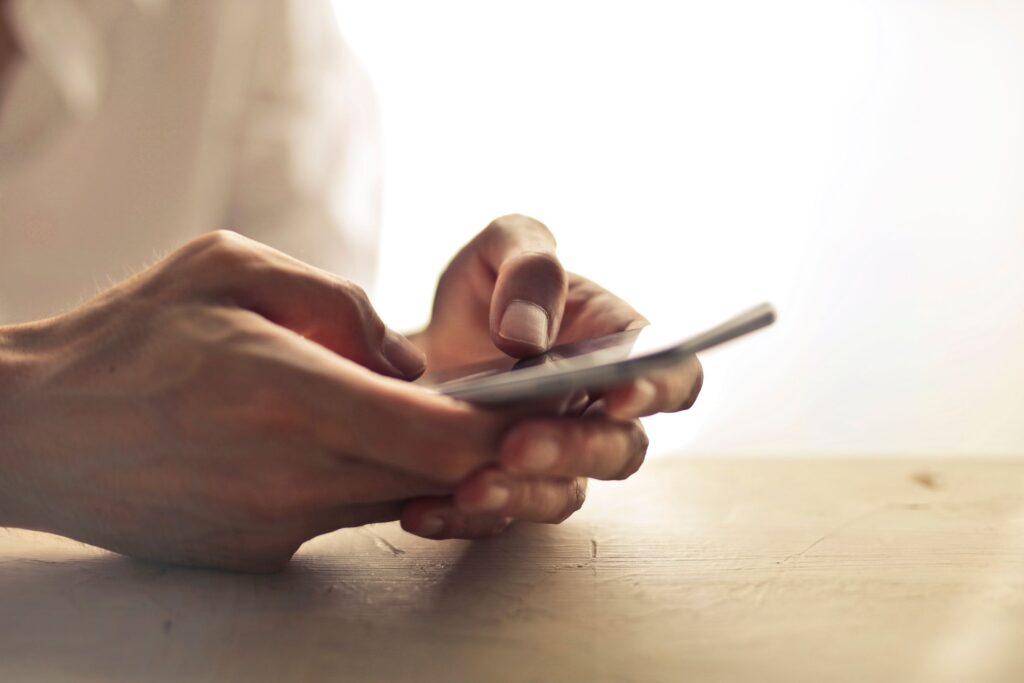 Close-up of a person's hands holding a smartphone at a wooden table in soft, warm window light.