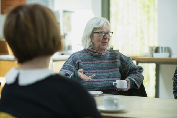 An older woman with white hair and glasses holds a cup of tea and talks with her hands. She is seen across a table, past the back of a younger woman's head.