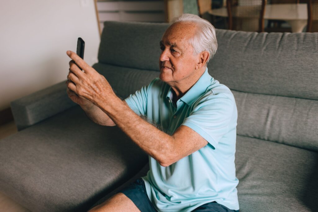 An older man sitting on a grey couch at home, holding his phone up at arm's length to record a message