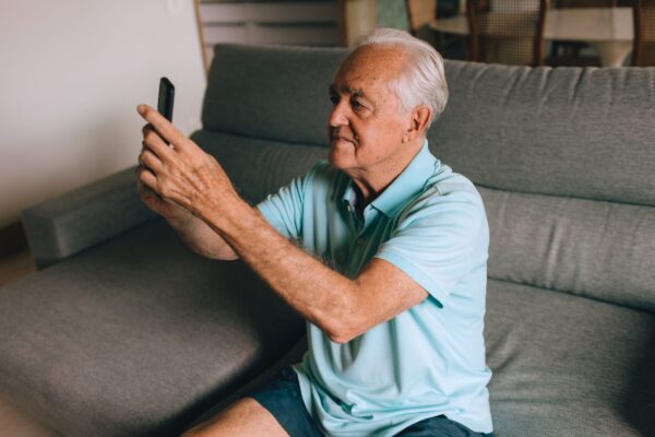 An older man sitting on a grey couch at home, holding his phone up at arm's length to record a message